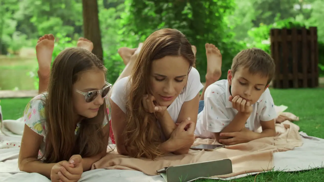 Woman with kids watching movie on pad Family lying on blanket in summer forest