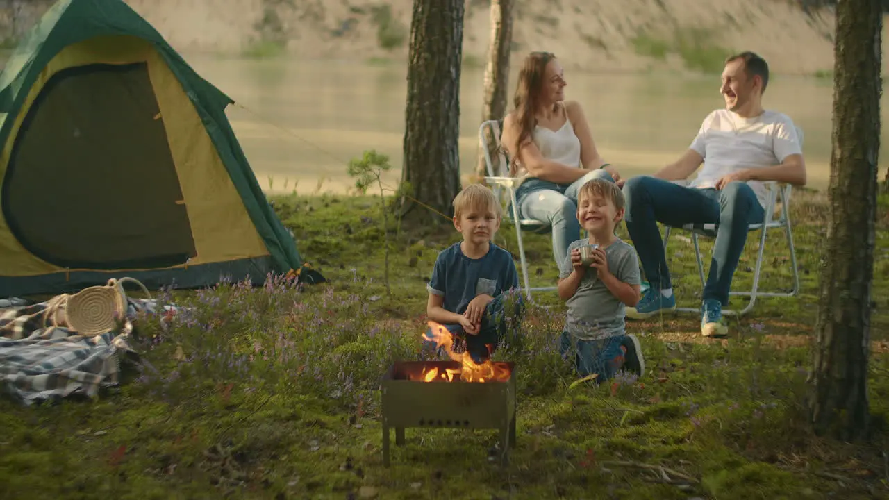 A family in nature parents watch as two boys at the fire roast marshmallows on sticks in the background of the tent Tent camp as a family