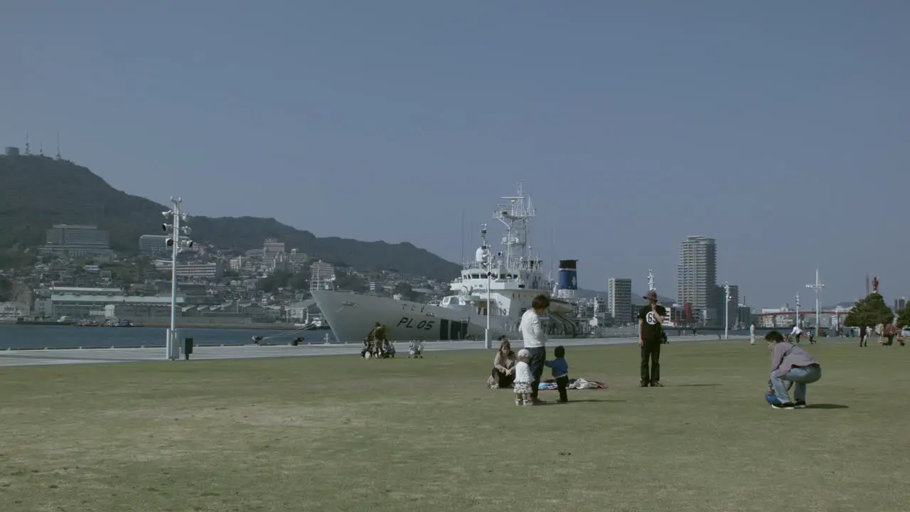 Family relaxing on green grass with a docked boat in Nagasaki port in the background Japan