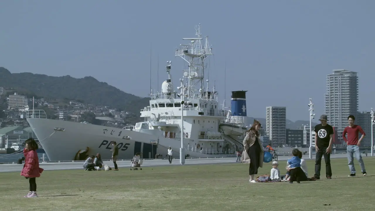 Group of adults and children on green grass with a docked boat in Nagasaki port in the background Japan