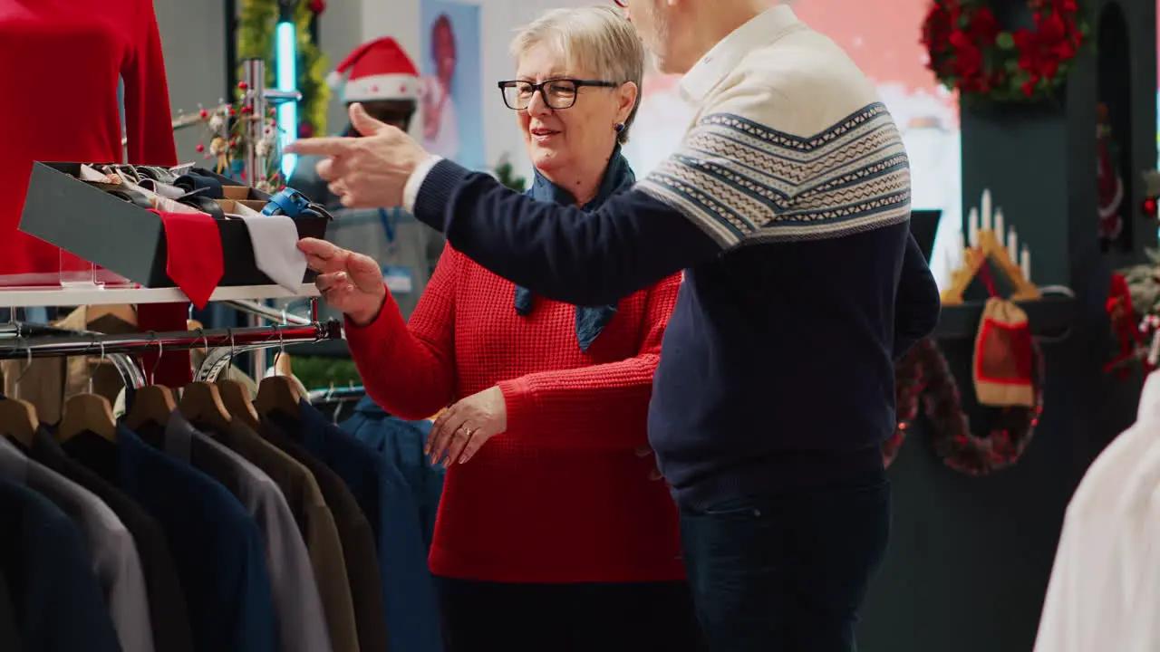 Woman browsing through xmas adorn clothing store racks with husband in Christmas shopping spree Elderly couple searching for elegant attire garments as gift for son during winter holiday season