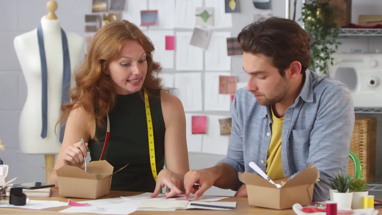 Male And Female Fashion Designers Have Working Takeaway Lunch Looking At Designs In Studio Together