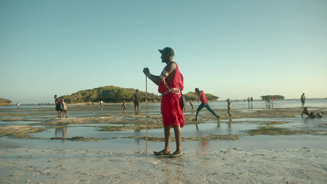 Masai Warrior Standing At The Beach With Locals Walking In Background On A Windy Day In Watamu Kenya