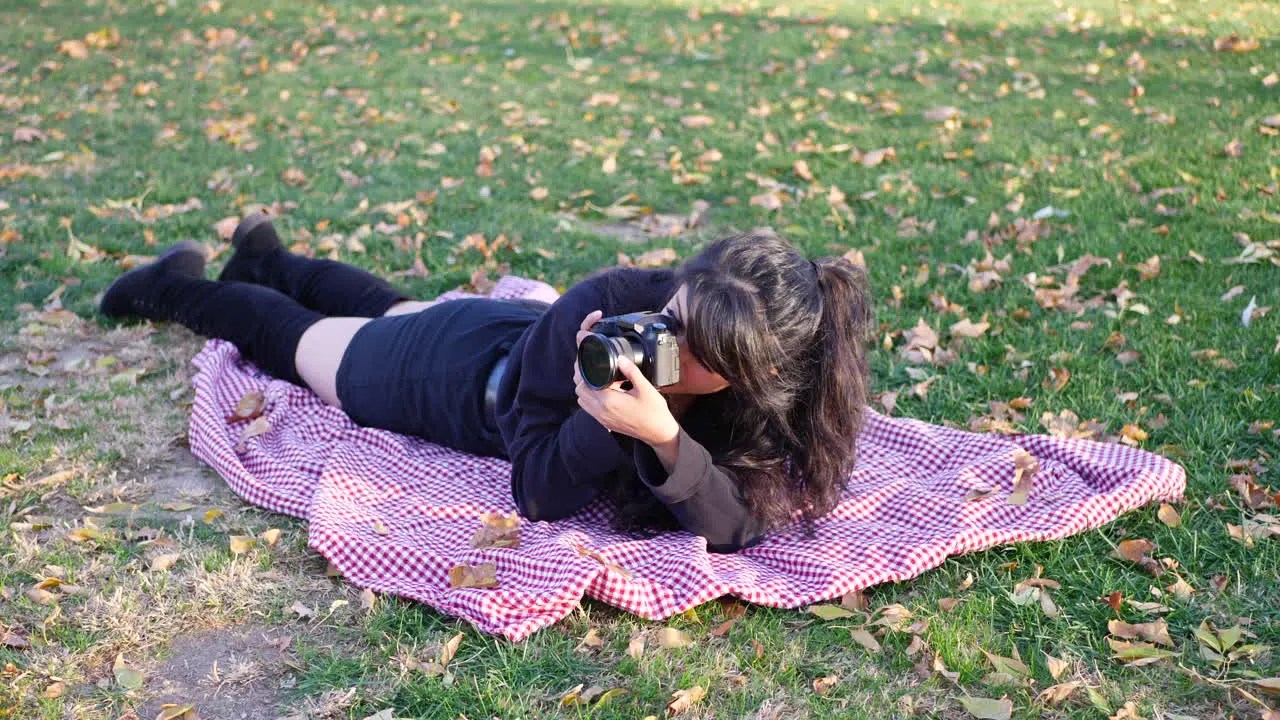 A young woman photographer taking pictures with her digital camera in a park during autumn with leaves all around the grass