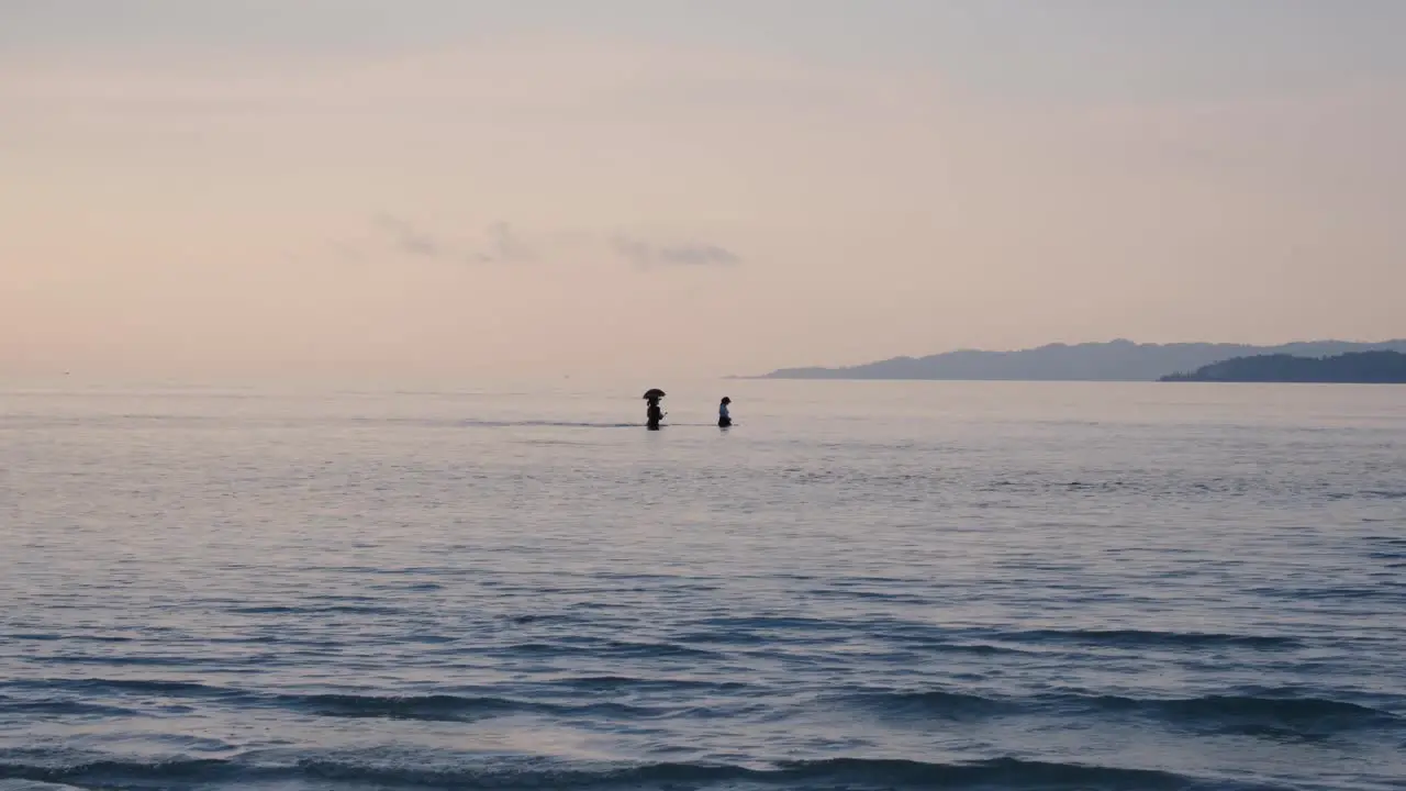 Small group of women with umbrella for sun protection walking across ocean lagoon between two tropical islands at low tide