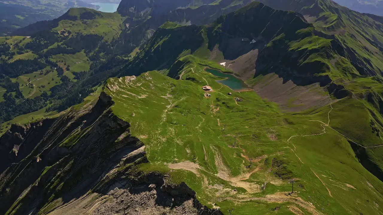 Aerial view of a mountain range in summer with green grass peaks and slopes and a little pond with a lake and mountains in the background