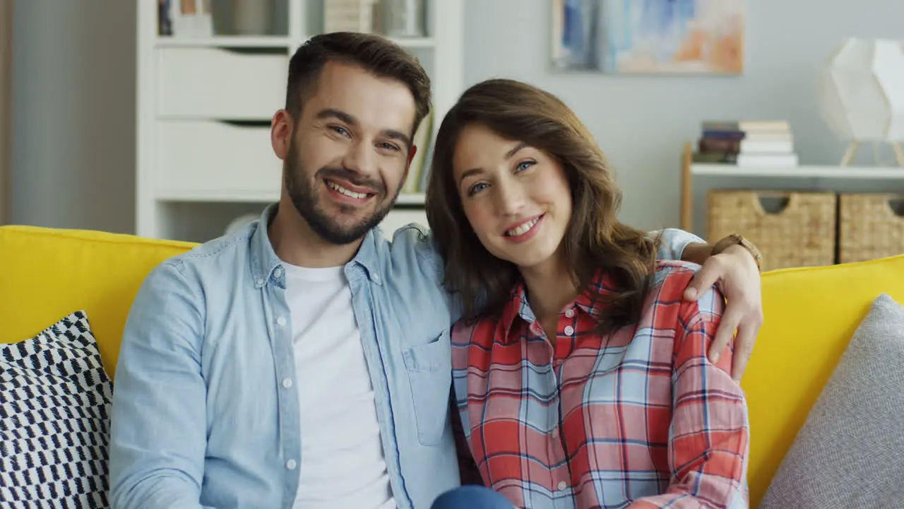 Portrait Shot Of The Young Smiled Boyfriend And Girlfriend Sitting On The Yellow Sofa And Posing To The Camera At Home