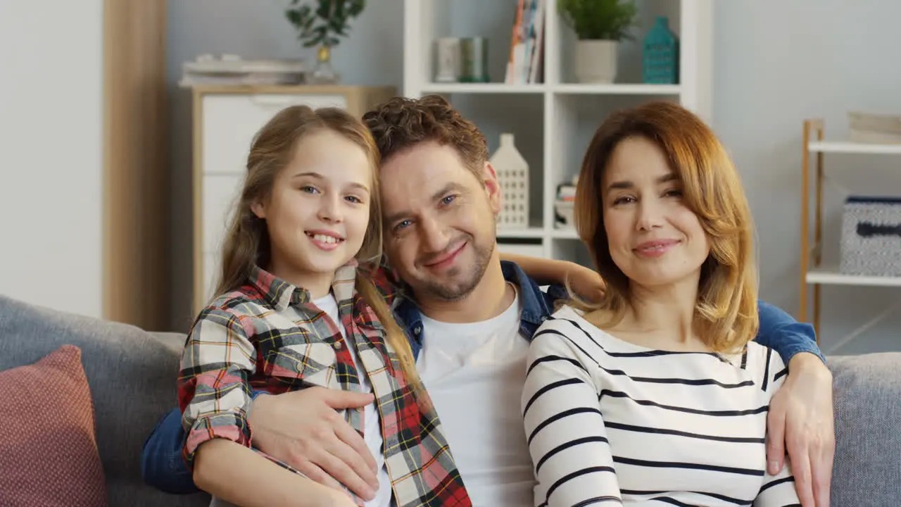 Portrait Of The Happy Good Looking Young Mother And Father Sitting On The Sofa At Home With Their Teenage Pretty Daughter Smiling And Posing To The Camera