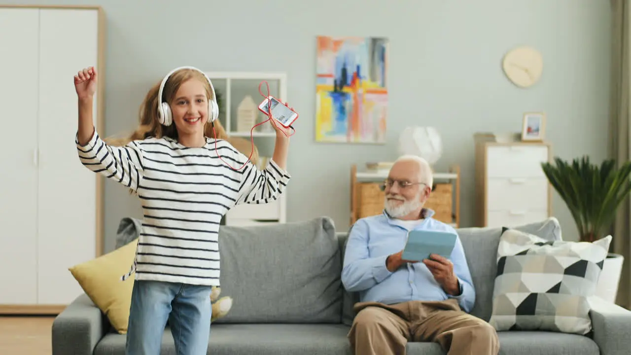 Pretty Funny Teenage Girl With Long Blond Hair And In The Headphones Listening To The Music On The Smartphone Dancing And Jumping In The Living Room While Her Grandpa Sitting On The Couch With Tablet Device