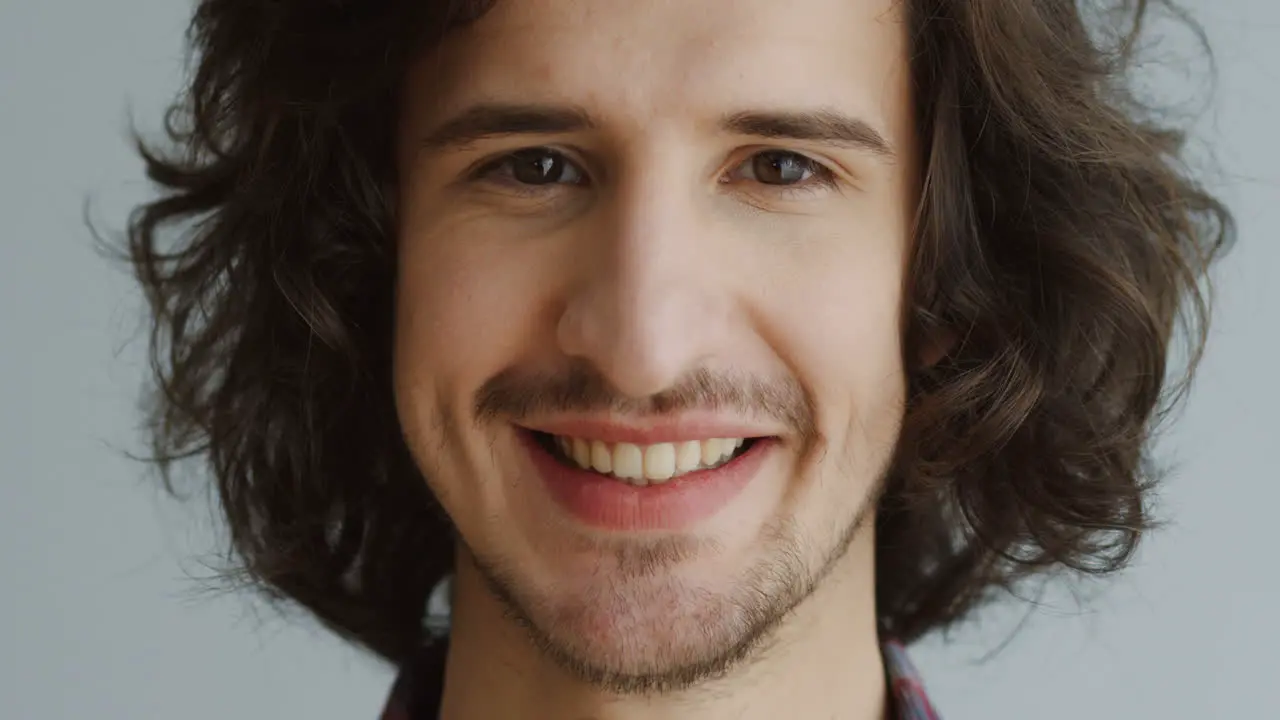 Close Up Of The Man's Face With Long Hair Looking Straight In The Camera And Smiling On The White Wall Background