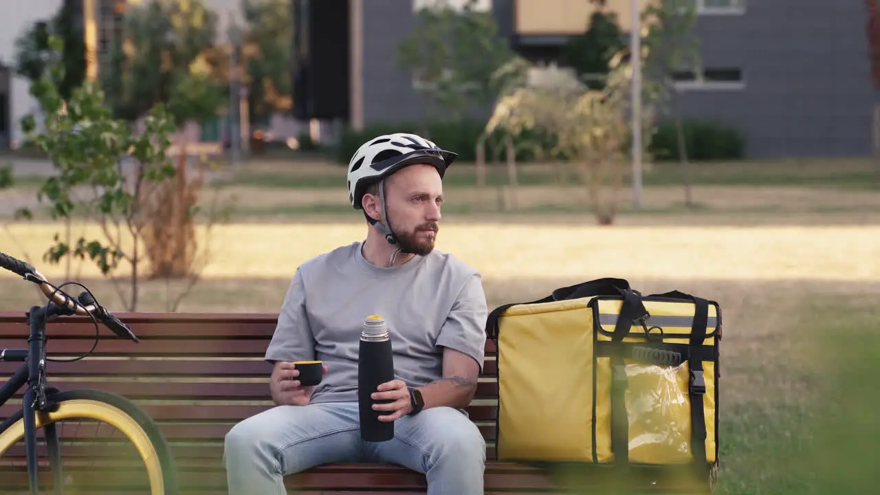 Tired Food Delivery Man Seated On A Bench Next To His Thermal Backpack During His Break Drinking A Cup Of Tea And Thinking About His Stuff