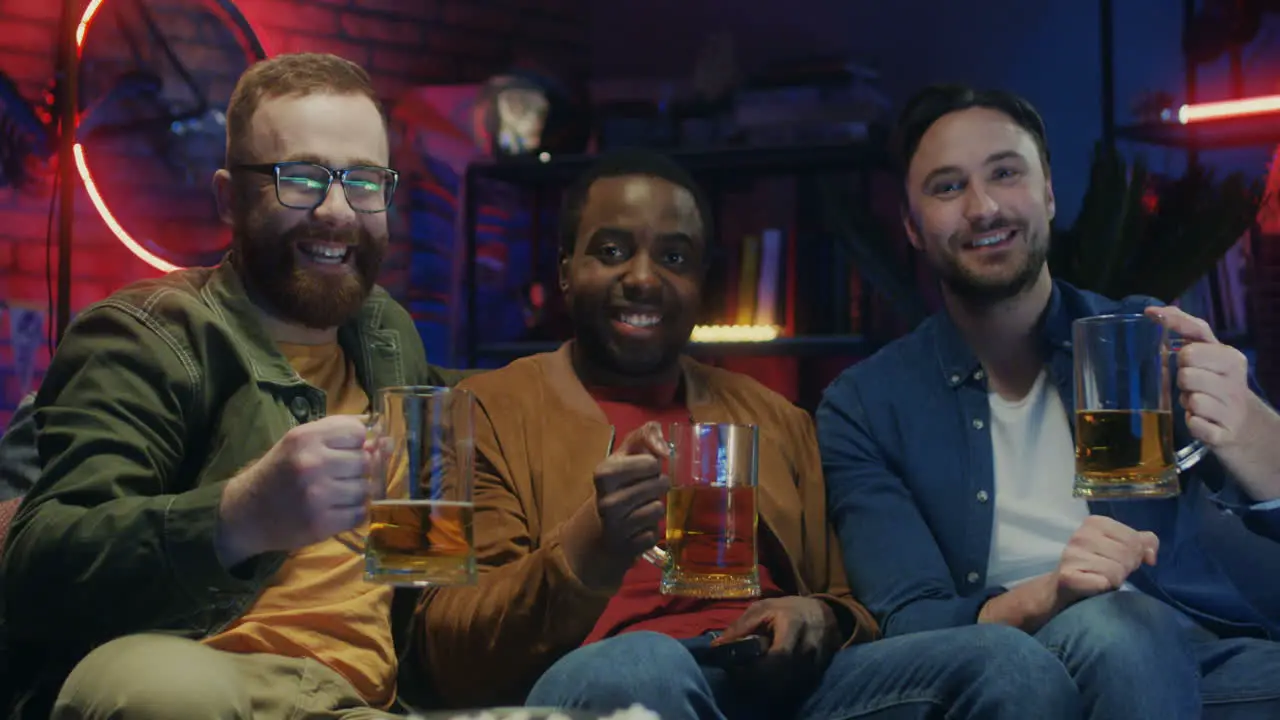 Three Multiethnic Young Guys Doing Cheers Gesture With Beer And Smiling To The Camera At Home At Night While Watching Tv