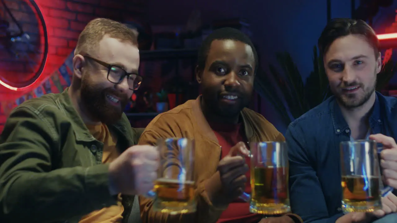 Mixed Races Young Three Men Doing Cheers Gesture With Beer And Smiling Cheerfully To The Camera In The Dark Room While Watching Sport Game On Tv