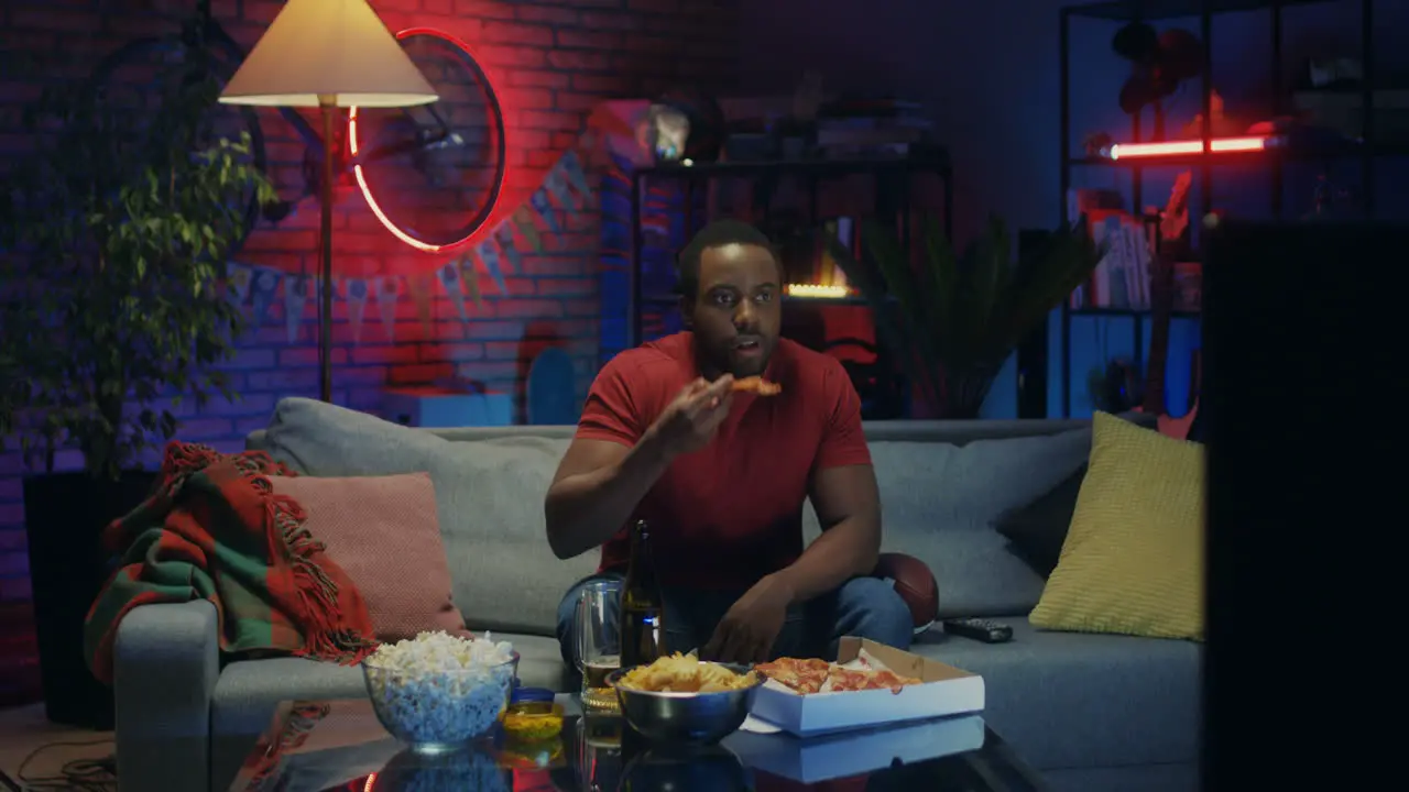 Young Attractive Man Sitting On The Sofa In The Living Room At Night Eating Pizza And Watching Tv