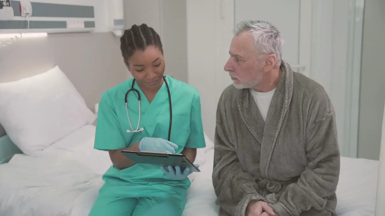 A Beautiful Young American Female Doctor Sitting On The Edge Of A Hospital Bed With An Old Patient