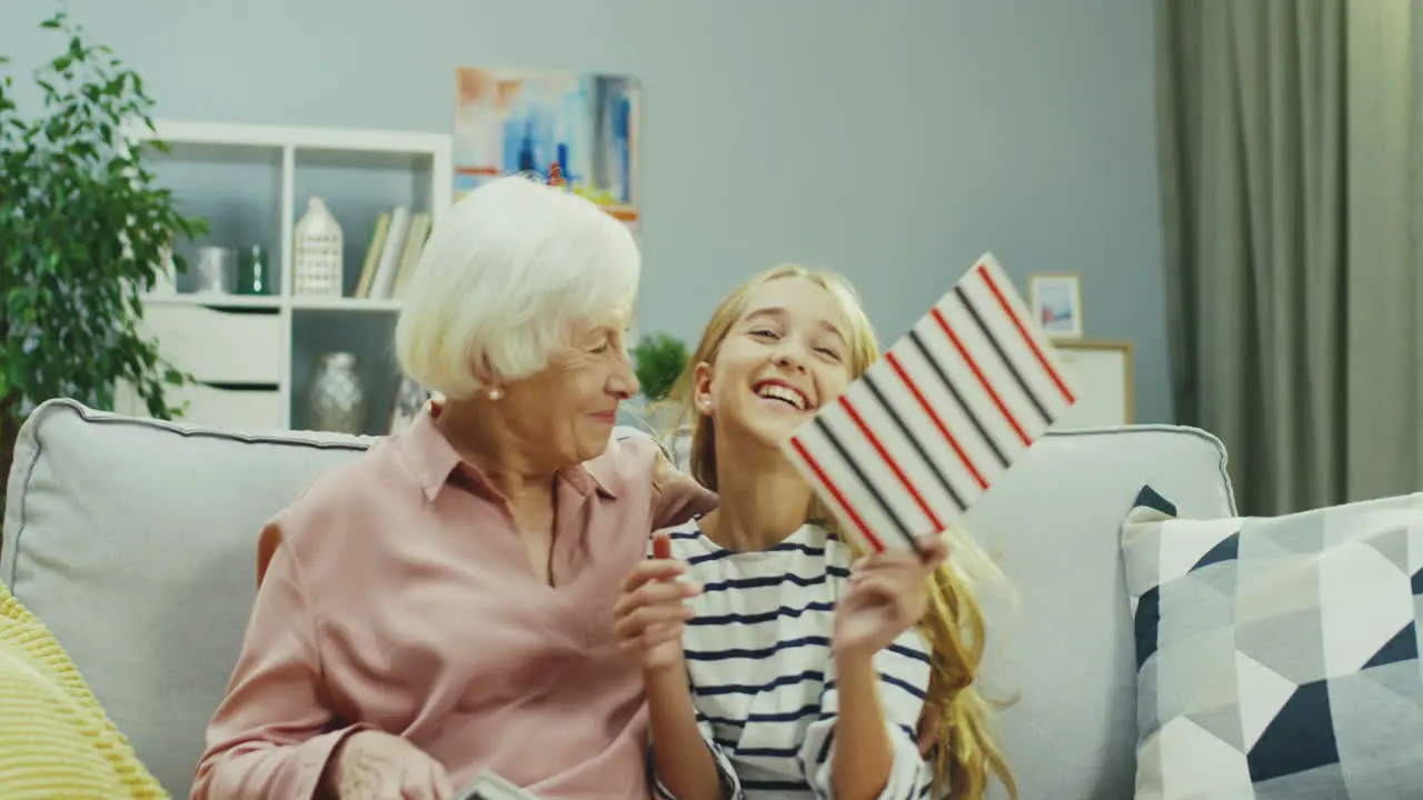 Grandmother Sitting On The Couch At Home While Her Cute Teenager Granddaughter Coming To Her With Copybook From School And Showing Off With Her Good Marks