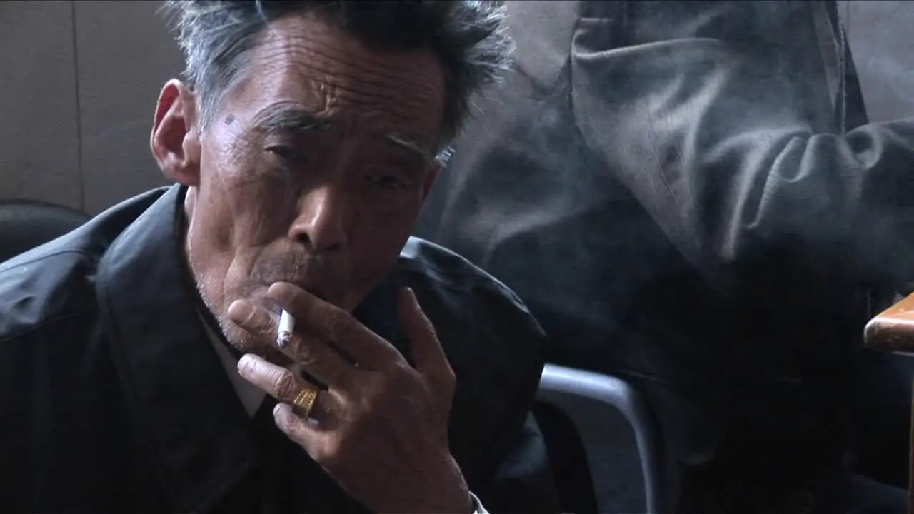 An old Chinese man smokes a cigarette and peers at the camera