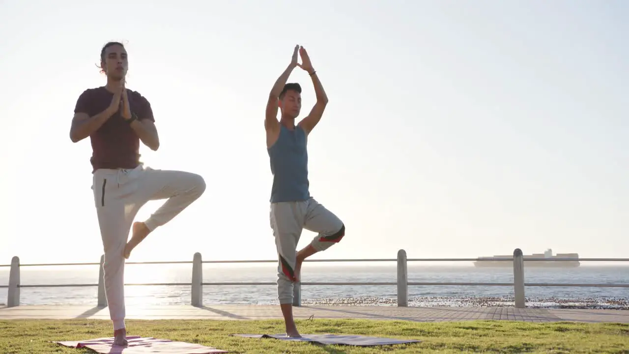 Happy diverse gay male couple doing yoga and meditating at promenade by the sea slow motion