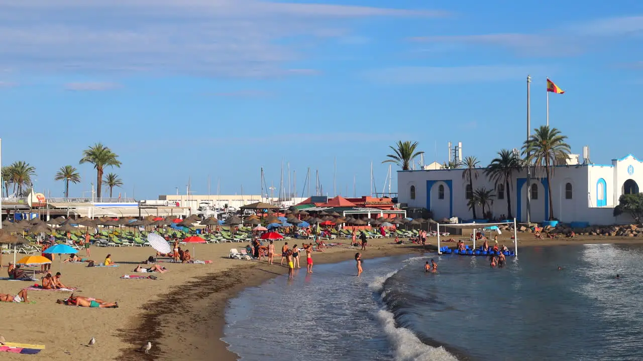 People enjoying a beautiful sunny beach day in Marbella port fun sea vacation holiday destination in Malaga Spain 4K static shot