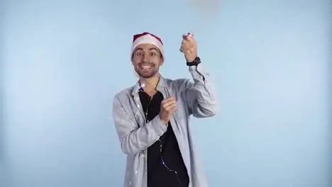 Young smiling man in casual clothes and santa hat on a head exploding confetti cracker on a blue background and starting to jump in excitement Happy celebrating of a New Year or Chrismas