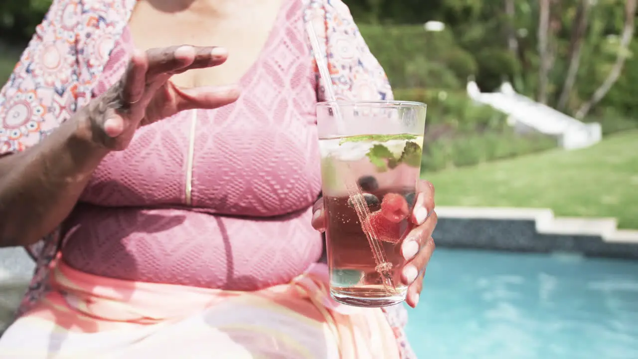 Happy senior african american woman in sunglasses by pool drinking cocktail slow motion