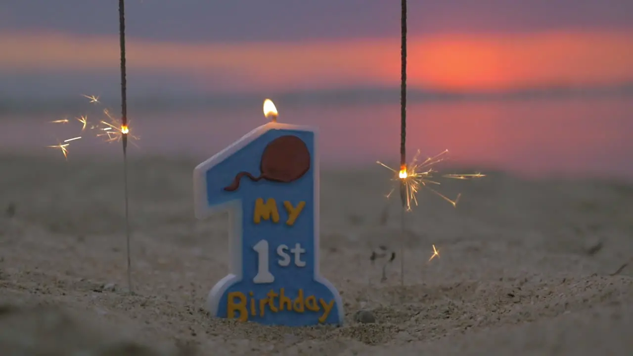 Slow motion view of candle and two sparklers standing in the sand on beach against blurred sunset