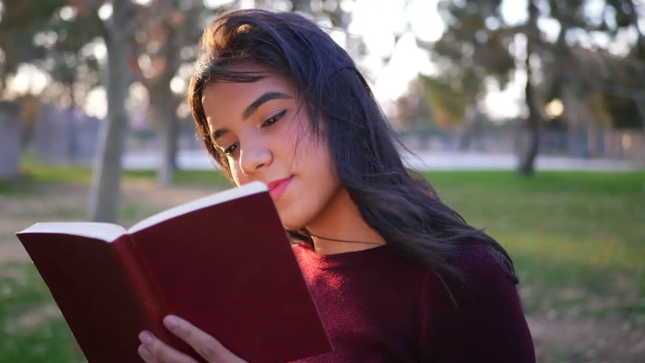 Close up on a young hispanic woman college student reading the pages of a red story book or novel outdoors at sunset SLOW MOTION