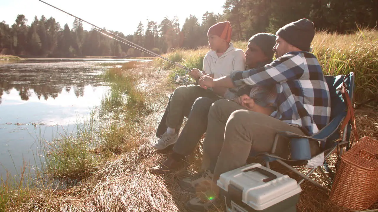 Grandfather and father sit showing boy how to fish by a lake