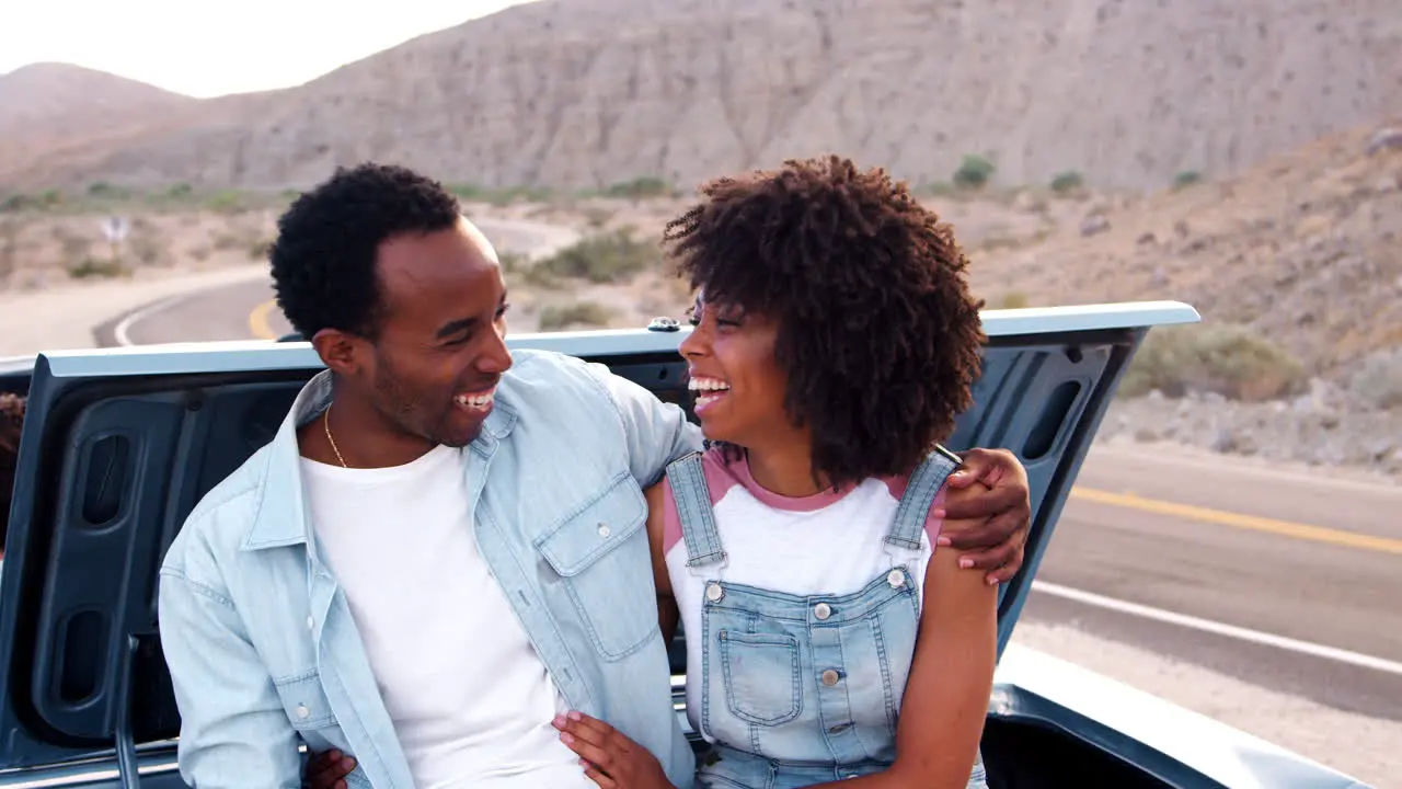 Laughing couple sitting on car at roadside stop on road trip