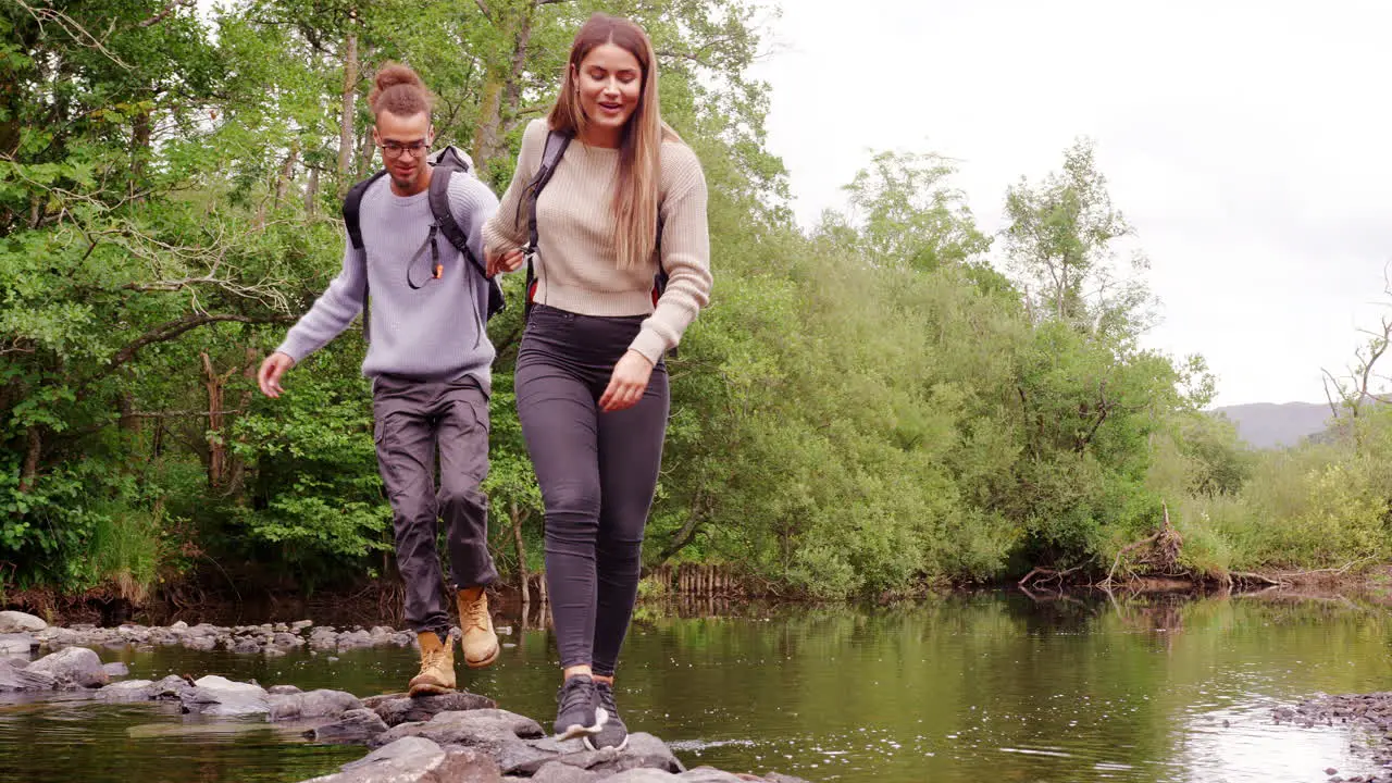 A young adult couple hold hands while crossing a stream balancing on stones during a hike