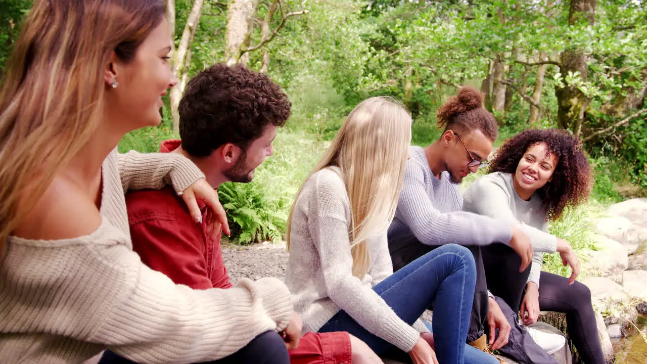 Five young adult friends taking a break during a hike sitting on rocks by a stream in a forest handheld