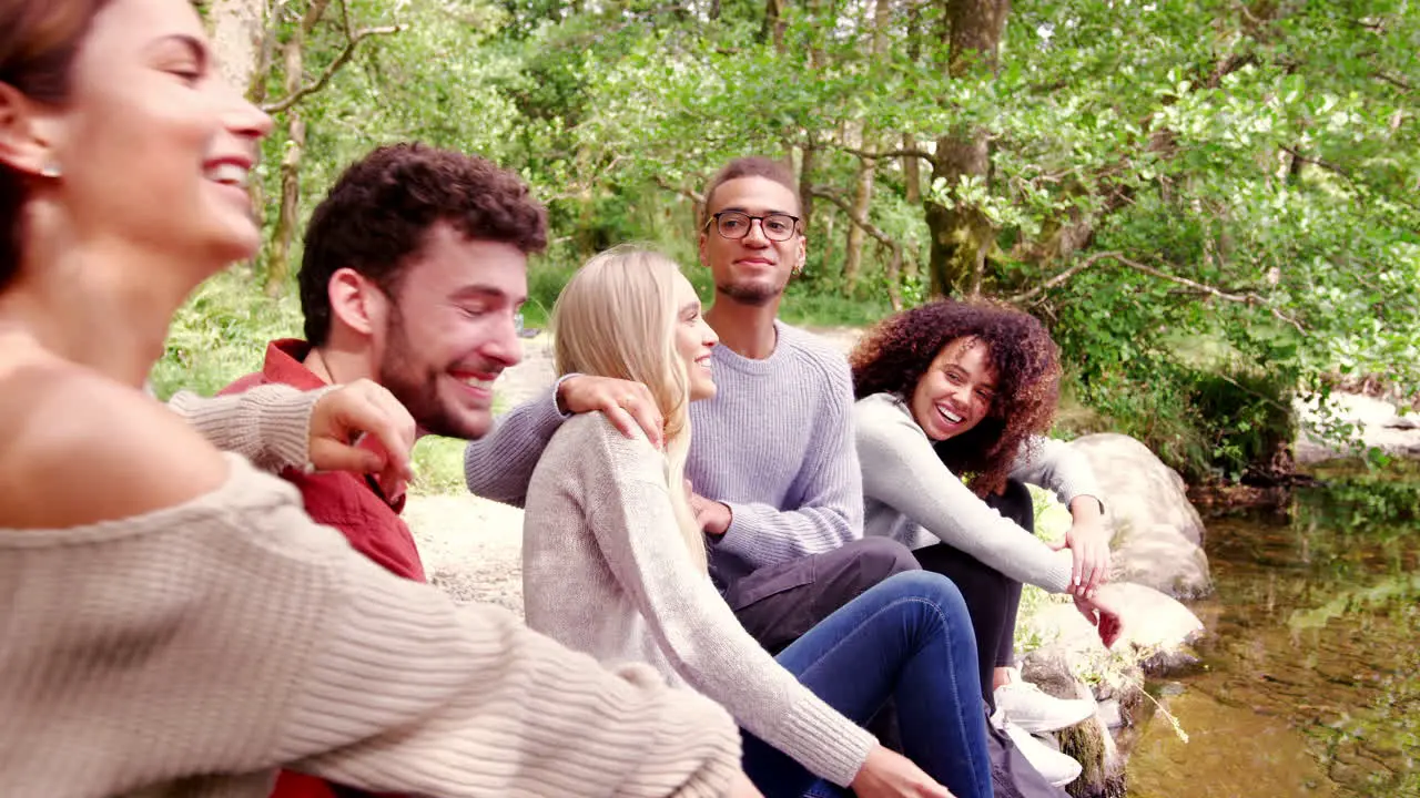 Five young adult friends taking a break during a hike sit talking by a stream in a forest handheld