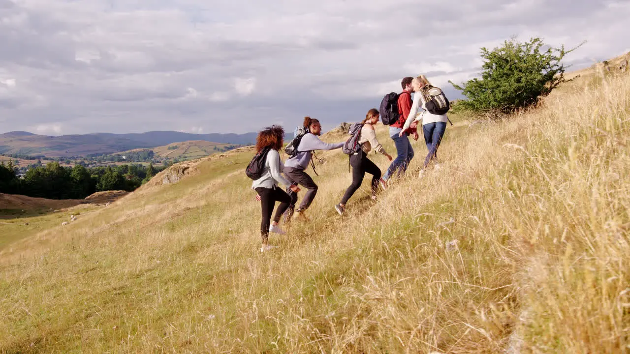 A multi ethnic group of five young adult friends help each other while hiking uphill across a field to the summit of a mountain tracking shot