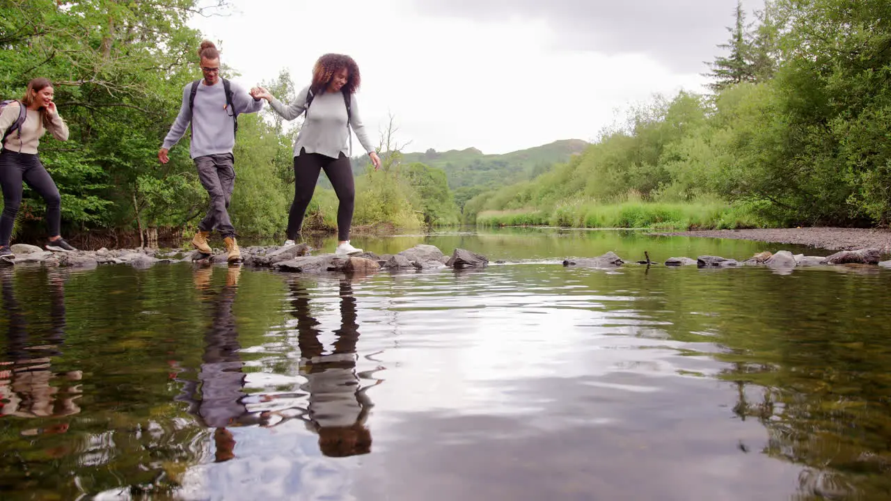 Five young adult friends hold hands carefully crossing a stream balancing on stones during a hike lockdown