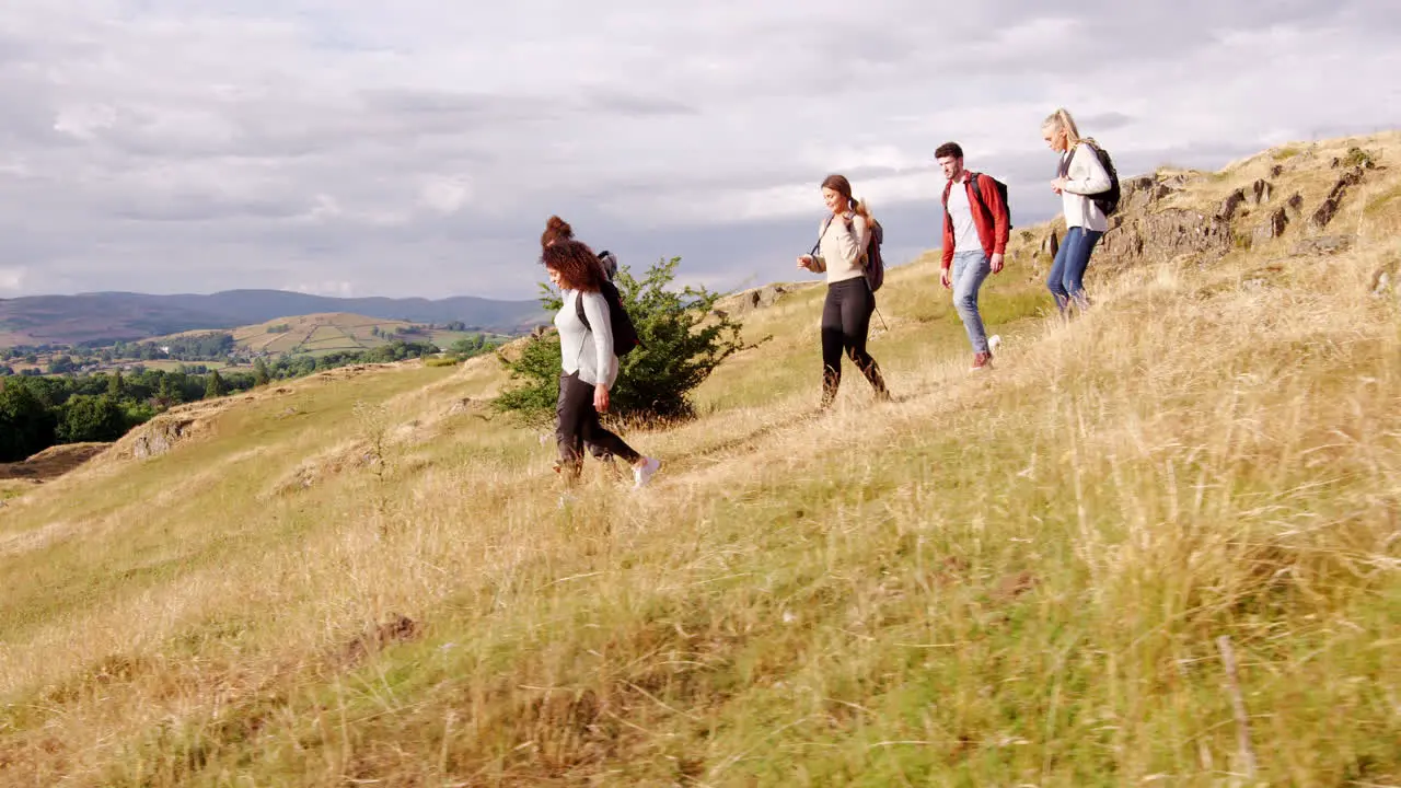 A multi ethnic group of smiling young adult friends help each other while walking downhill in a field during a mountain hike