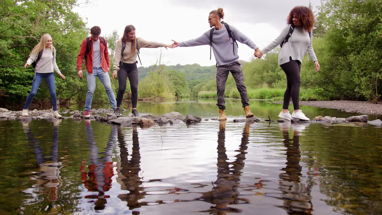 Five young adult friends hold hands and help each other while crossing a stream balancing on stones during a hike lockdown