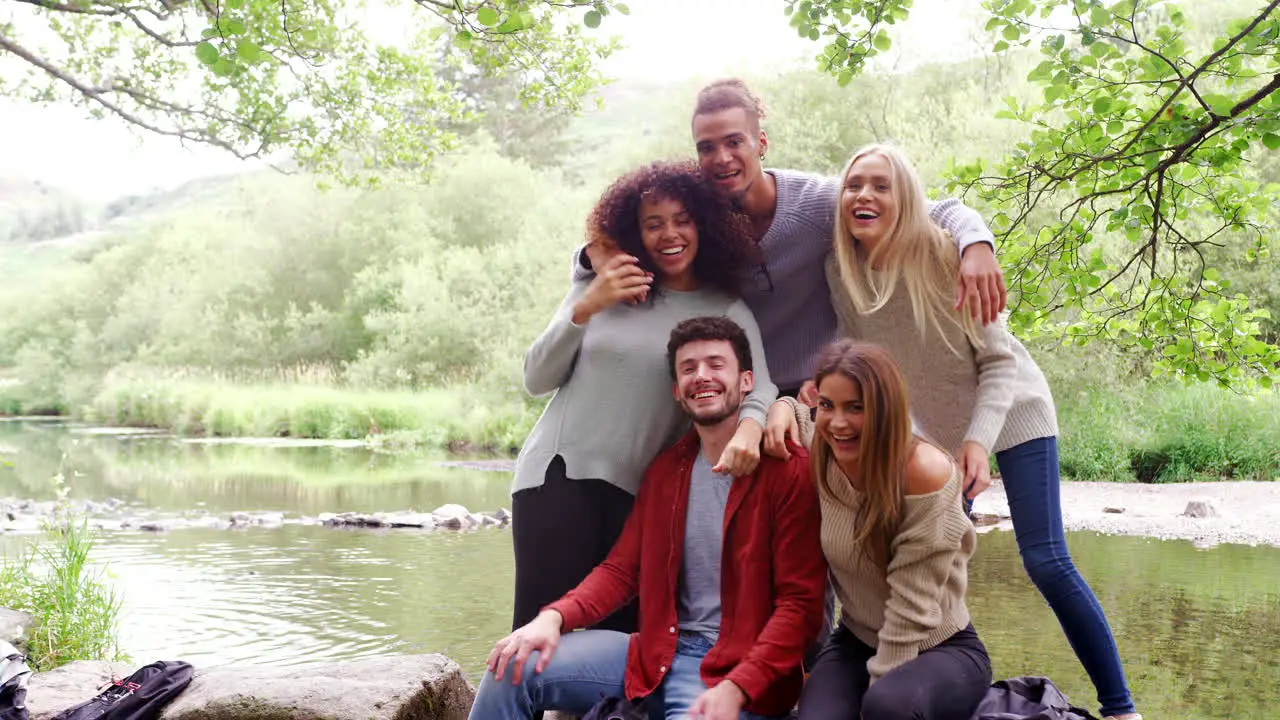 Five young adult friends take a break and pose to camera standing and sitting on rocks by a river during a hike handheld