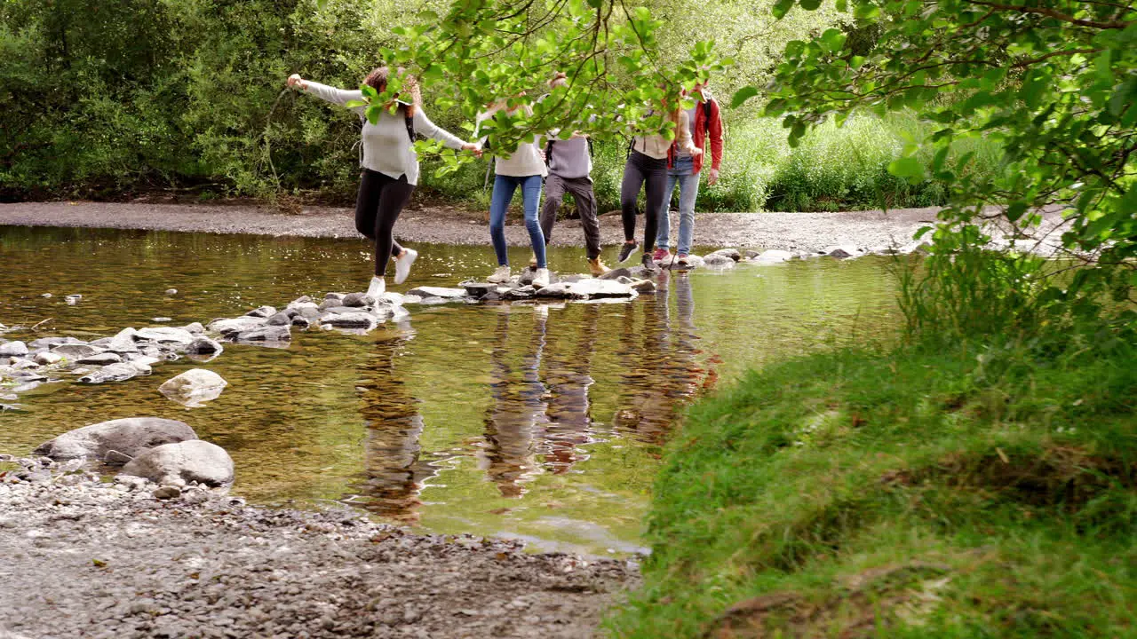 Five young adult friends hold hands and help each other cross a stream balancing on stones during a hike