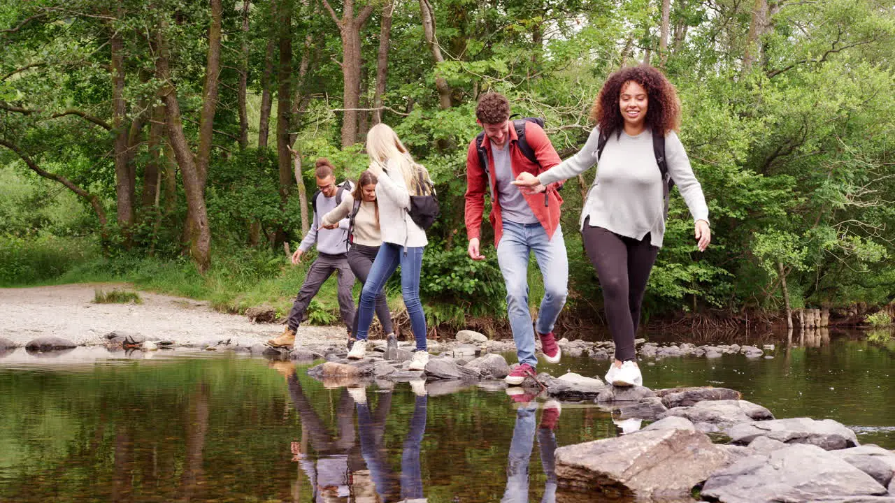 Five young adult friends hold hands and help each other while crossing a stream and standing on stones during a hike