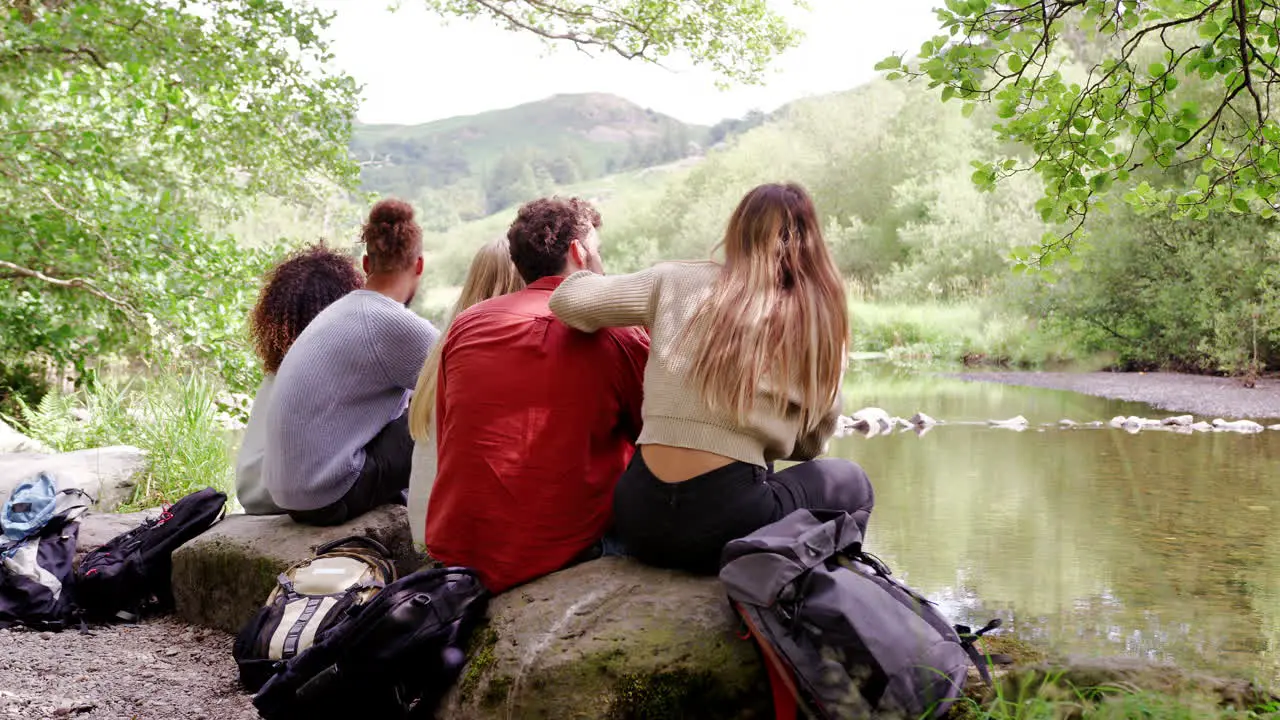 Five young adult friends admire the landscape while taking a break sitting on rocks by a river during a hike back view