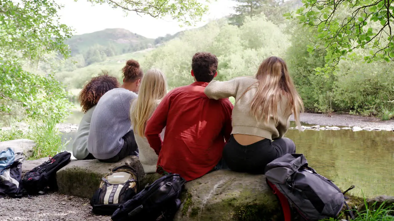 Five young adult friends admire the landscape while taking a break sitting on rocks by a stream during a hike back view