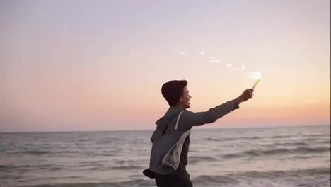 Young handsome african guy running by the sea during sunset and holding burning sparkling candle in his hand Slow Motion shot