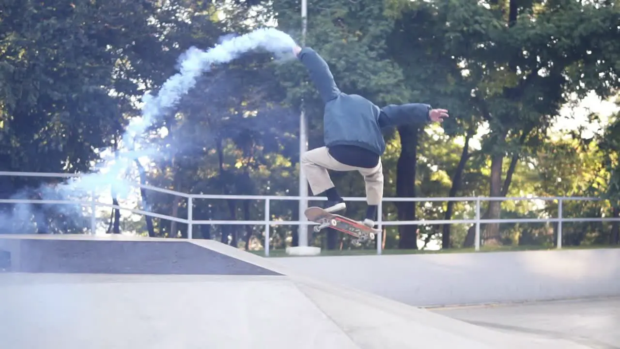 Young Guy Practicing On Sunny Summer Day While Holding Blue Coloured Burning Smoke Bomb On The Skate Ramp