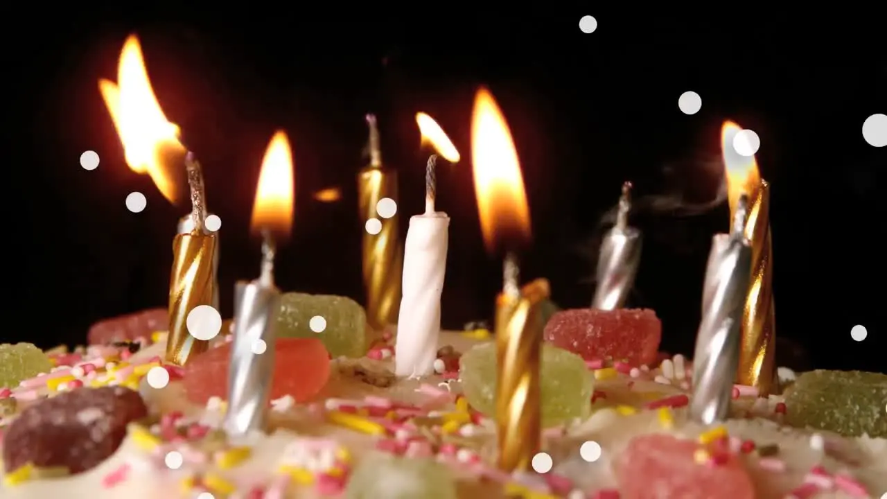 Animation of white spots floating over burning candles on a cake against black background