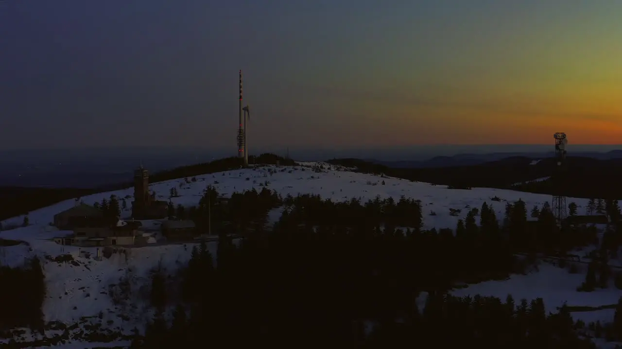 Aerial shot at dawn ascending over snow covered hill showing broadcasting tower wind turbine and buildings