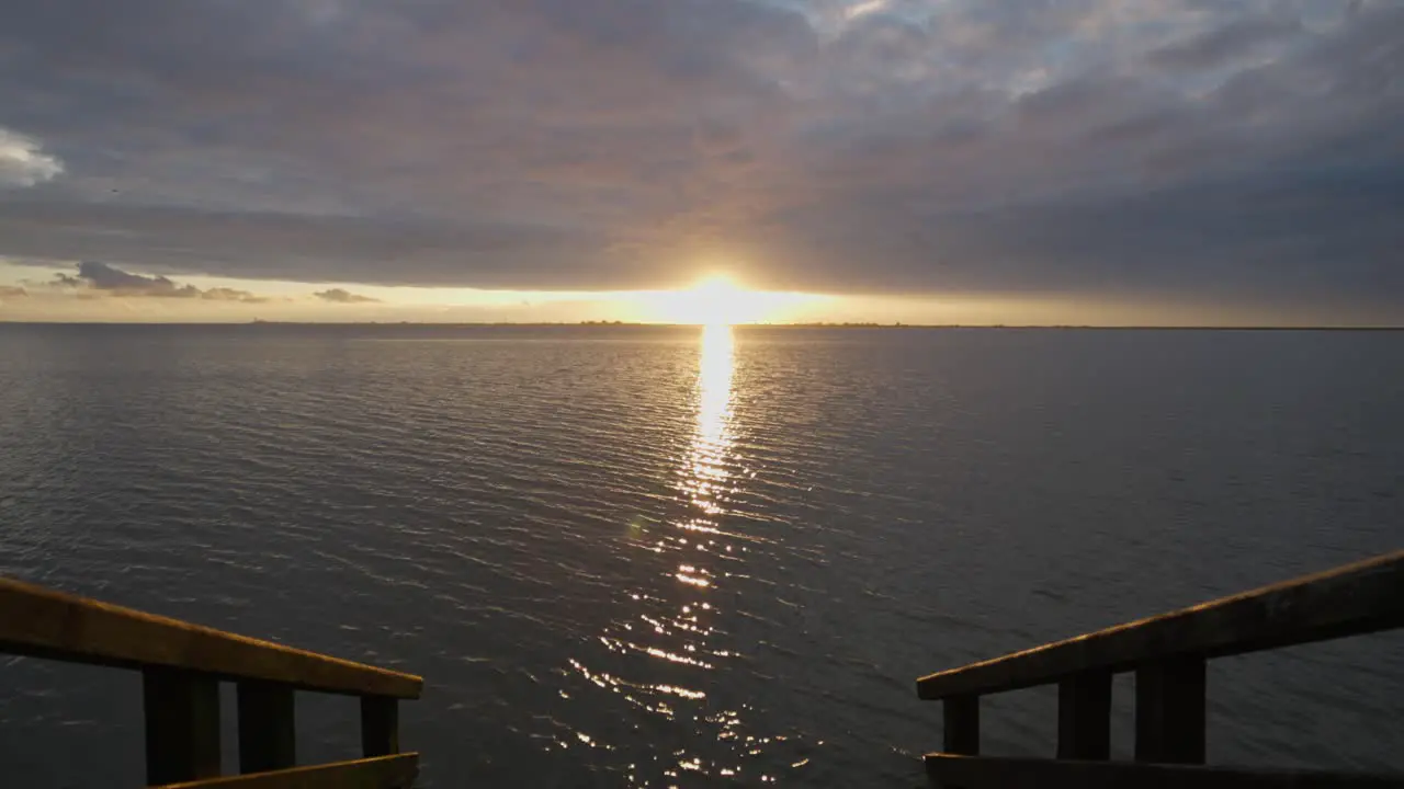 Tilt up from wooden stairs leading into north sea to golden sun over horizon seafront sunset sky with dramatic dense clouds