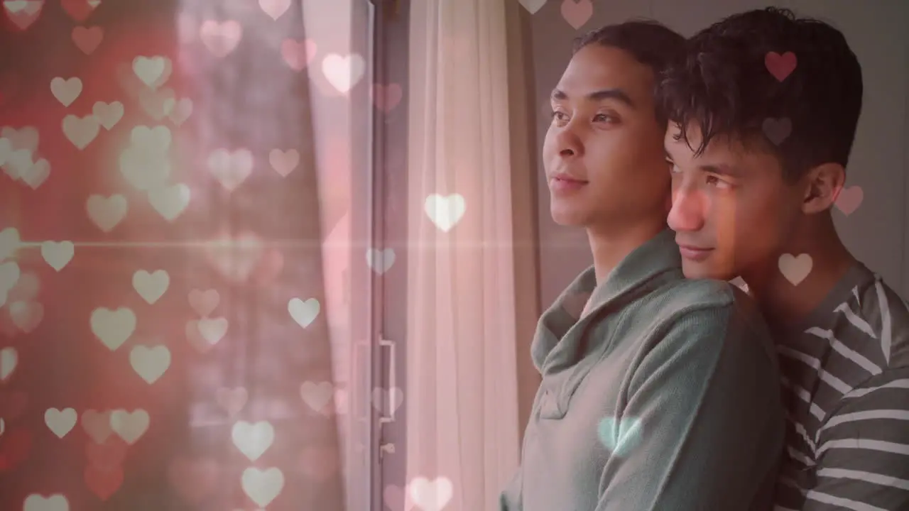 Animation of glittering hearts over happy diverse gay male couple looking out of window embracing