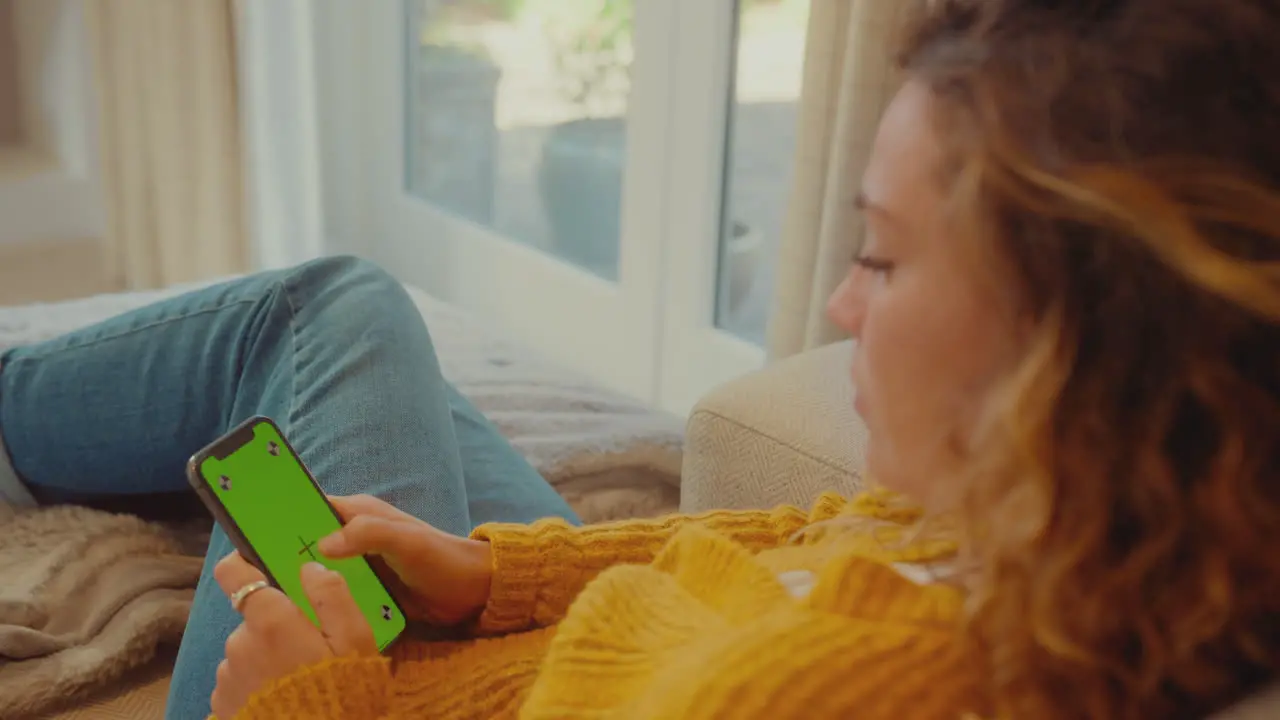 Over The Shoulder View Of Young Woman At Home Using Mobile Phone With Green Screen