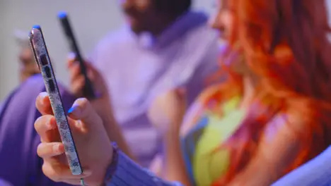 Close Up Studio Shot Of Young Gen Z Friends Sitting On Sofa Talking And Sharing Social Media Post On Mobile Phones 10