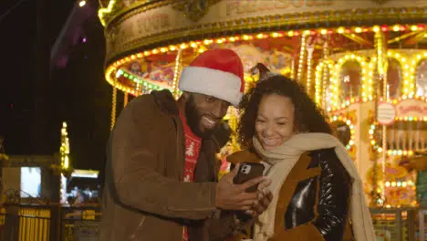 Couple Celebrating Christmas Standing By Fairground Roundabout On London South Bank At Night With Mobile Phone 1